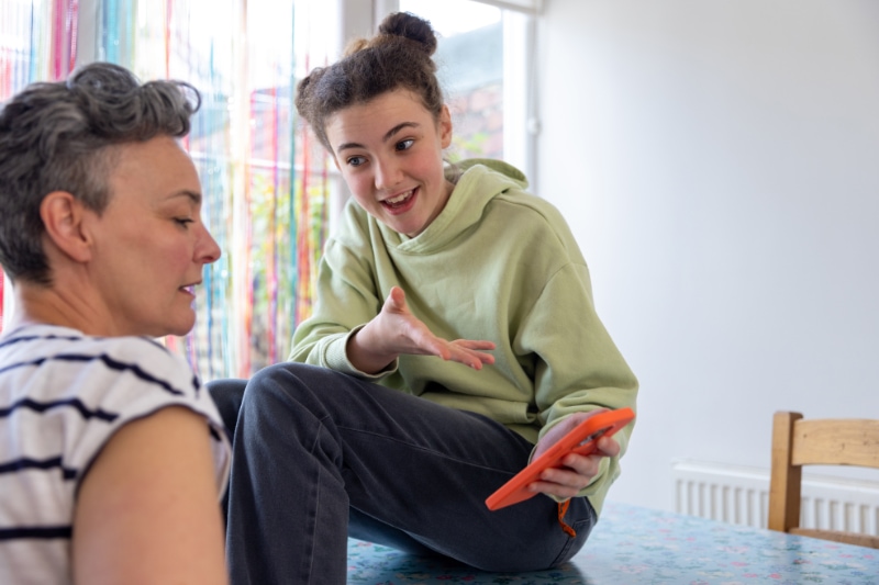 5 Common AC Problems. Over the shoulder shot of mother and daughter talking in the kitchen. The daughter is showing her mother something on an orange mobile phone.